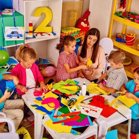 Mai multi copii care lucrează la măsuțe, alături de educatoare